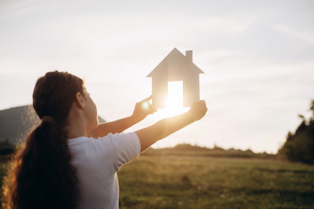 Gros plan d'une préadolescente tenant une maquette de maison en papier, sur fond de magnifique coucher de soleil et de montagnes majestueuses. Maison baignée de soleil dans la nature estivale. Maison de rêve. Concept de logement et de prêt immobilier.